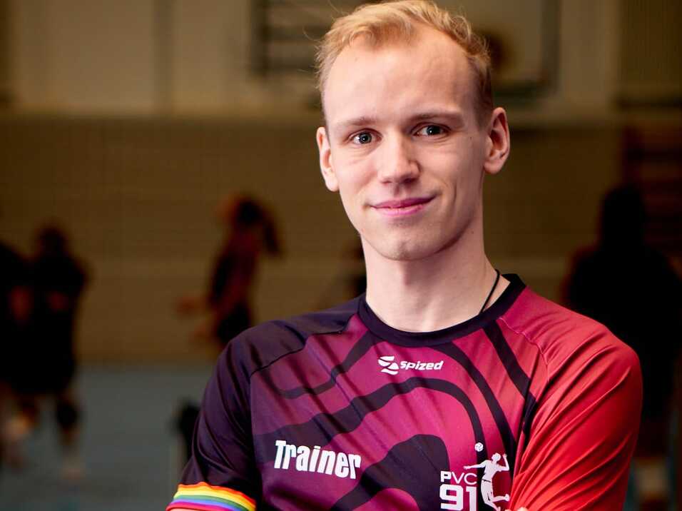 L&auml;chelnder Trainer in einem sportlichen Outfit mit &bdquo;PVC 91&ldquo;-Aufdruck, steht mit verschr&auml;nkten Armen auf einem Hallenboden.