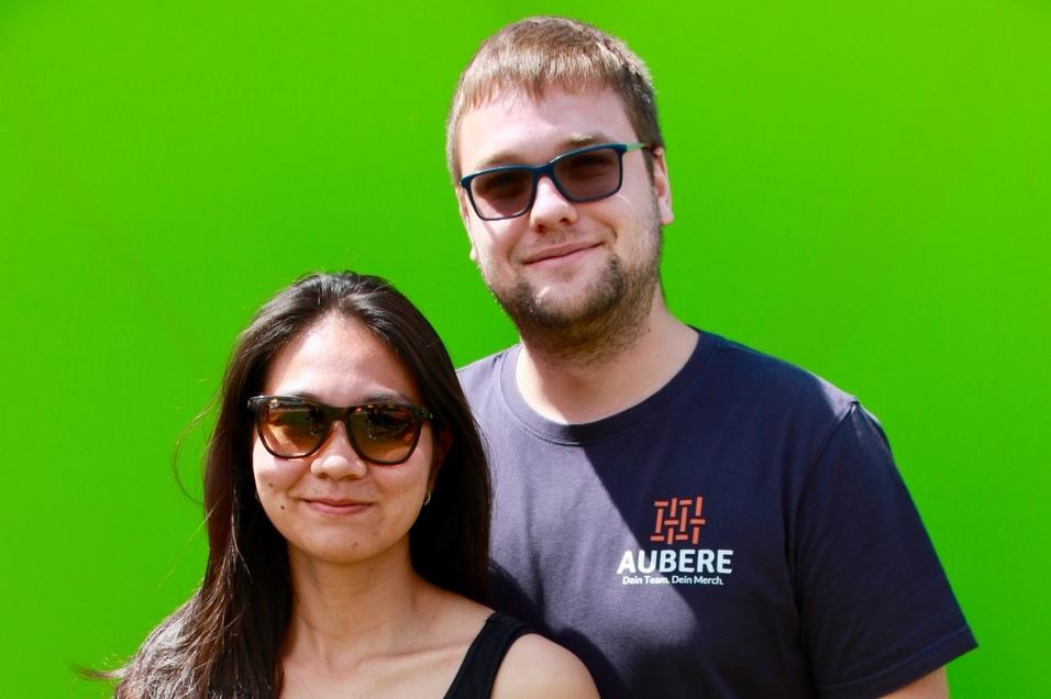 Paar steht vor einem grünen Hintergrund. Die Frau trägt ein schwarzes Top und der Mann ein dunkles T-Shirt.