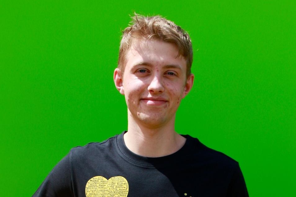 Junger Mann mit blondem Haar hält einen bunten Fußball, stehend vor einem grünen Hintergrund. Er trägt ein schwarzes T-Shirt.
