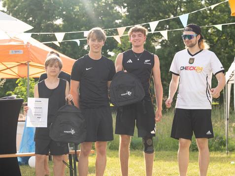 vier Jugendliche in Sportkleidung stehen lächelnd, während sie schwarze Rucksäcke in der Hand halten.