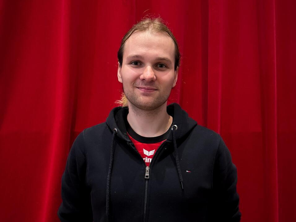 Junger Mann mit l&auml;chelndem Gesicht steht vor einem roten Vorhang, tr&auml;gt schwarzen Hoodie und T-Shirt.