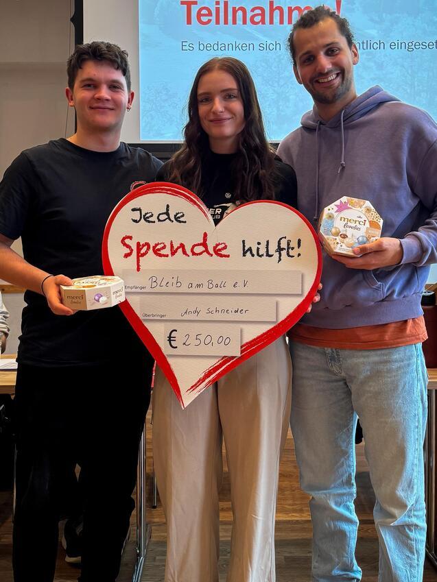 Drei Personen mit einer gro&szlig;en Herz-Form, beschriftet mit &bdquo;Jede Spende hilft!&ldquo;, stehen im Vordergrund eines Veranstaltungsraums.