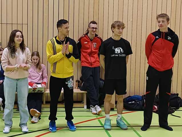 Gruppierung von fünf Personen in Sportkleidung, versammelt in einer Sporthalle, mit weiteren Personen im Hintergrund.