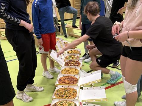 Gruppe von Jugendlichen um einen Tisch mit mehreren Pizzakartons in einer Sporthalle versammelt.