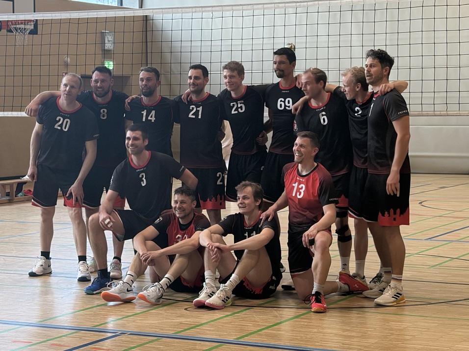 Gruppenschnitt von elf Volleyballspielern in schwarz-roten Trikots in einer Sporthalle vor einem Volleyballnetz.