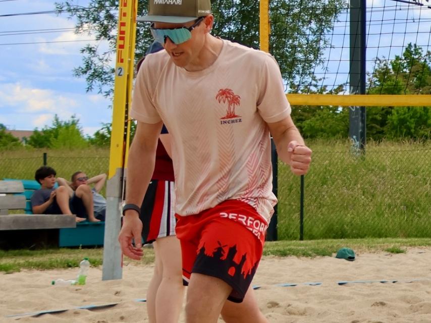 Sandplatz mit Spielern beim Beachvolleyball, einer hat einen Ball in der Hand, andere laufen oder stehen.