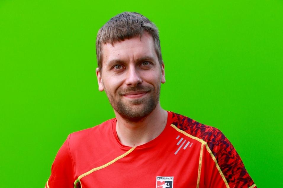 Mann mit kurzem Haar in rotem Sportshirt posiert vor grünem Hintergrund, Hände auf den Hüften.