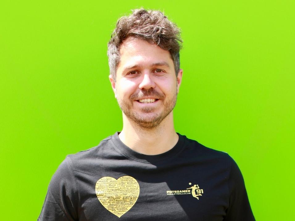 Mann mit lockigem Haar in schwarzem T-Shirt h&auml;lt einen Volleyball vor einer gr&uuml;nen Wand.
