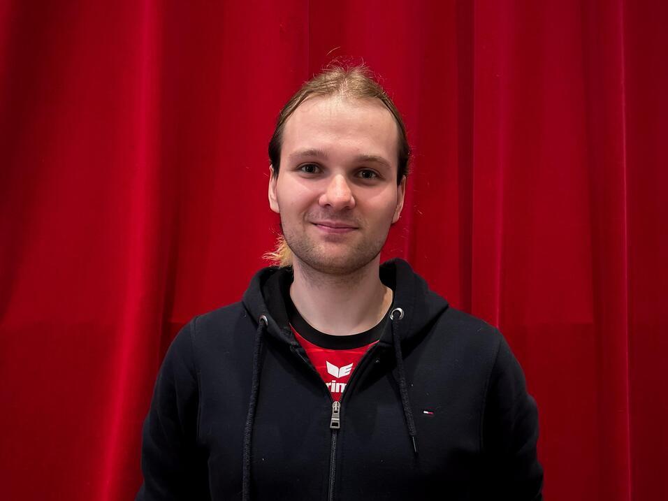 Junger Mann mit l&auml;chelndem Gesicht steht vor einem roten Vorhang, tr&auml;gt schwarzen Hoodie und T-Shirt.