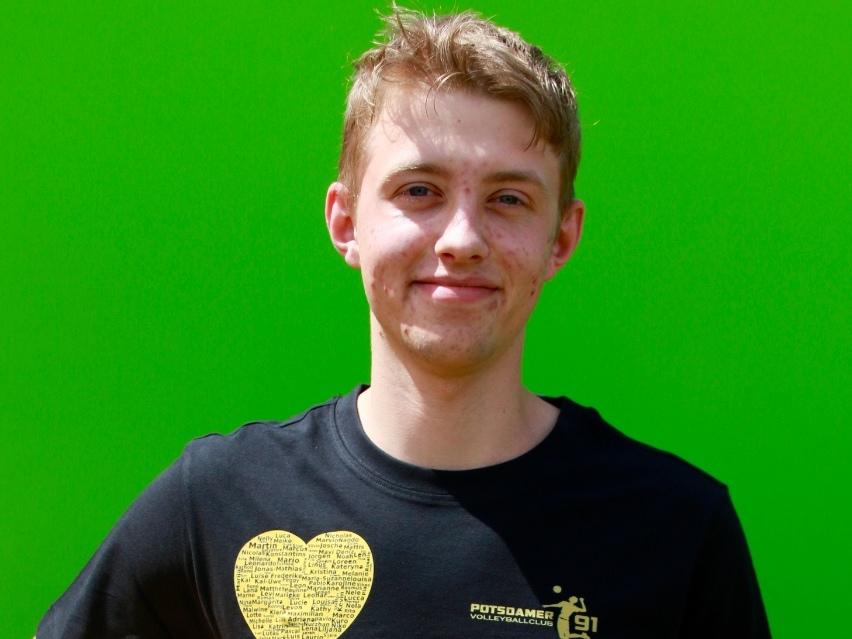 Junger Mann mit blondem Haar hält einen bunten Fußball, stehend vor einem grünen Hintergrund. Er trägt ein schwarzes T-Shirt.