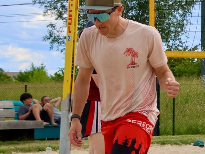 Sandplatz mit Spielern beim Beachvolleyball, einer hat einen Ball in der Hand, andere laufen oder stehen.