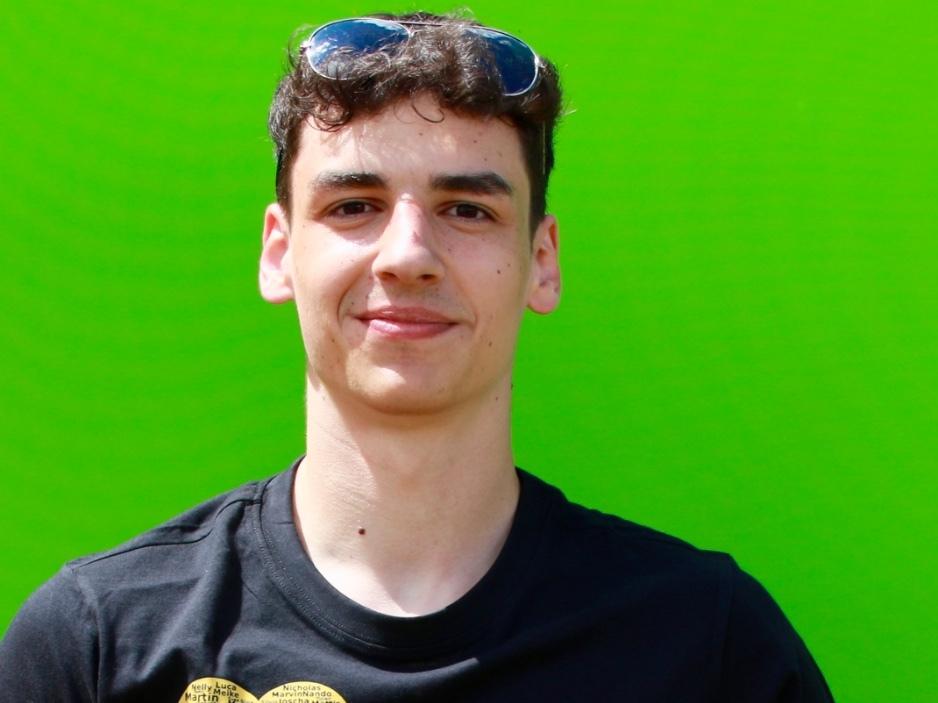 Junger Mann mit Sonnenbrille in schwarzem T-Shirt mit goldenem Herz und Volleyball vor gr&uuml;ner Wand.