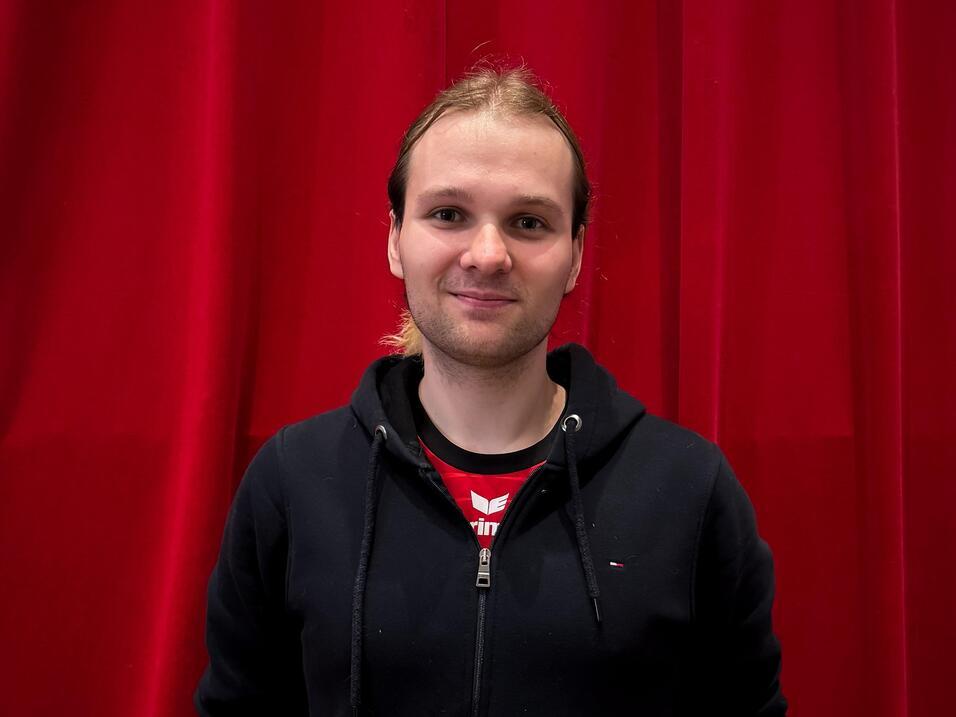 Junger Mann mit lächelndem Gesicht steht vor einem roten Vorhang, trägt schwarzen Hoodie und T-Shirt.
