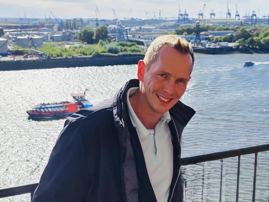Lächelnder Mann steht an einem Geländer mit Blick auf einen Hafen und Wasser bei sonnigem Wetter.