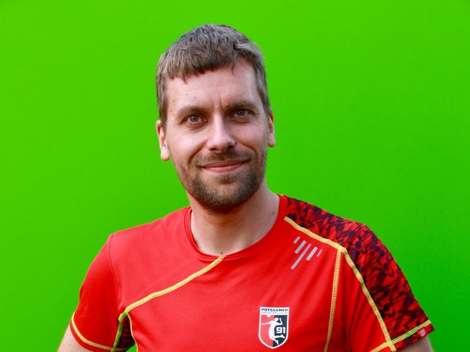 Mann mit kurzem Haar in rotem Sportshirt posiert vor grünem Hintergrund, Hände auf den Hüften.