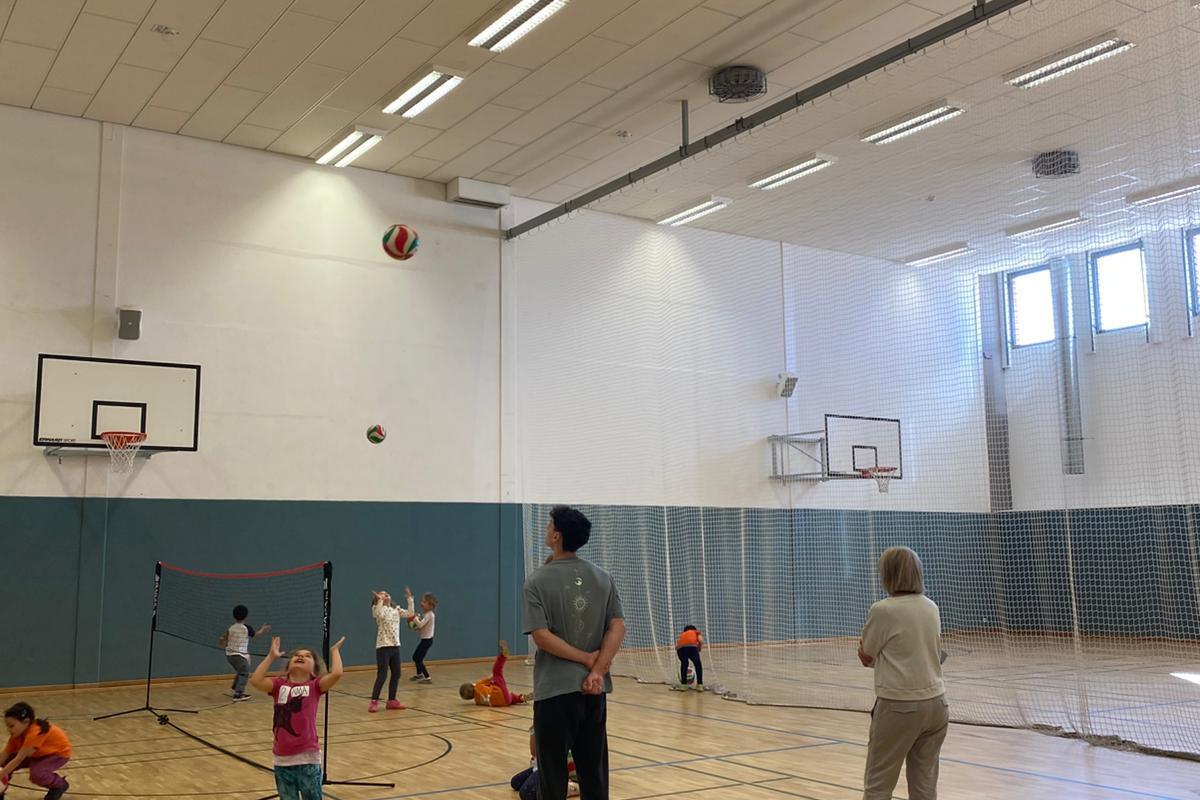 Kinder und Erwachsene spielen in einer Sporthalle mit einem Ball, während ein Netz und Basketballkörbe im Hintergrund sichtbar sind.