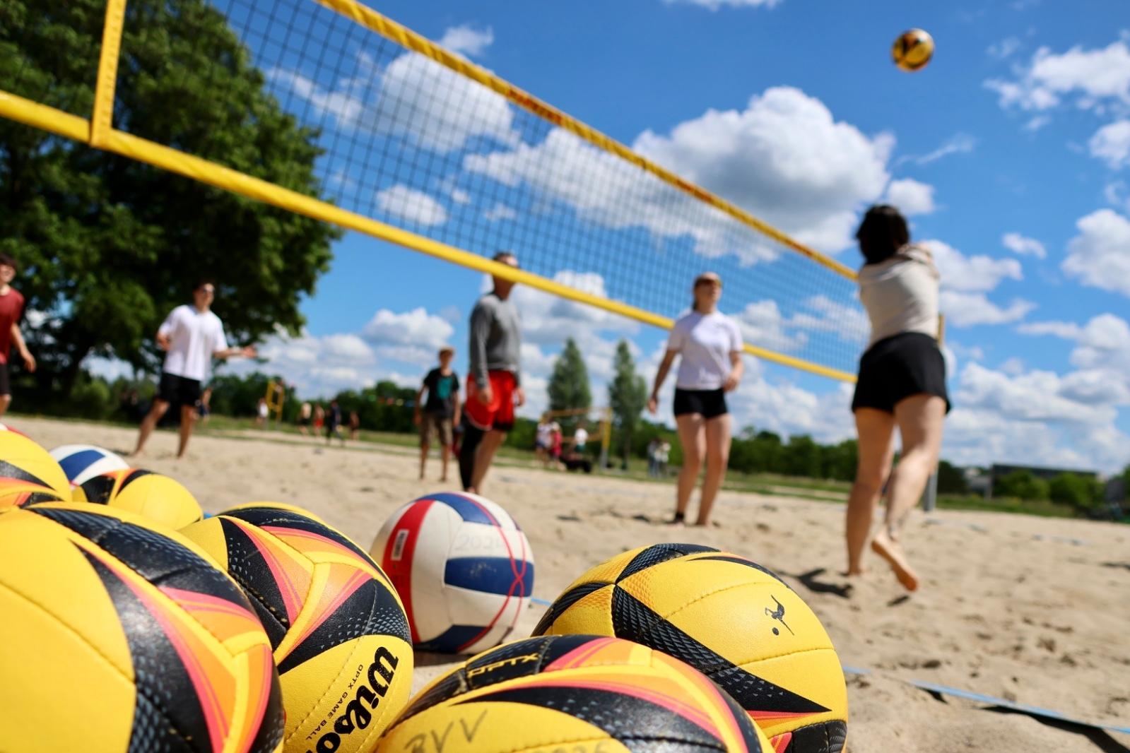 Nahaufnahme mehrerer Beachvolleyballs im Sand, mit Spielern im Hintergrund, die am Netz gegeneinander spielen.