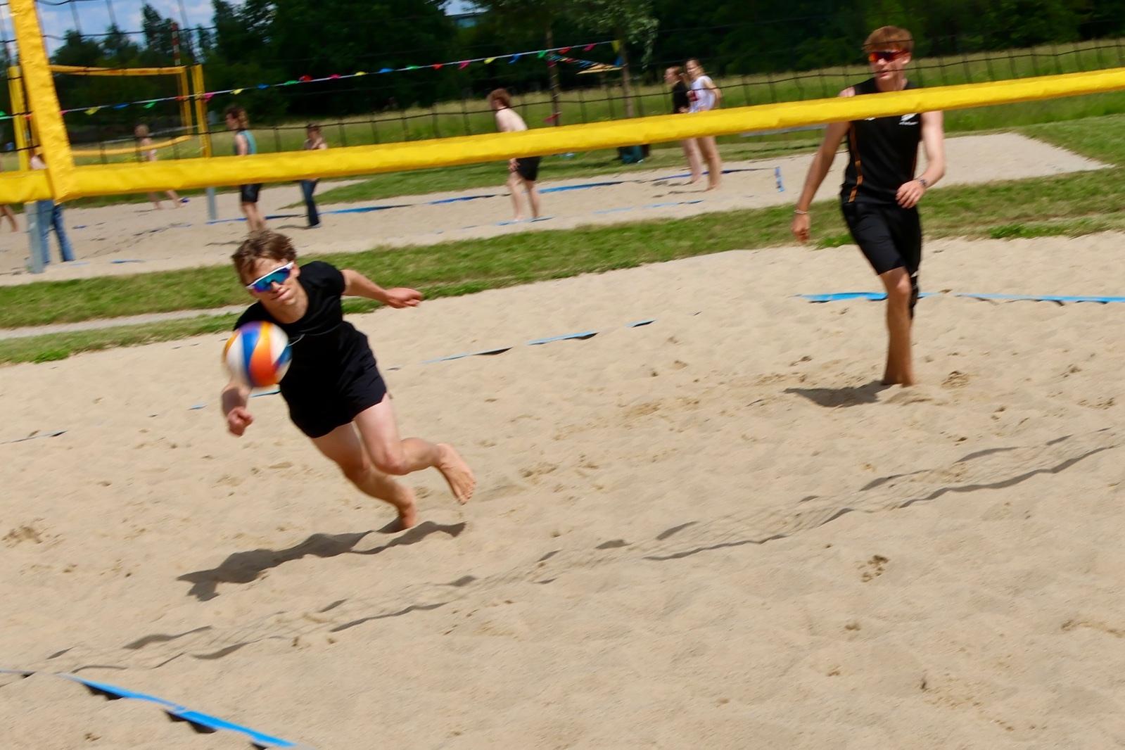 Zwei Jugendliche spielen Beachvolleyball auf einem Sandplatz; einer springt, um den Ball zu erreichen.