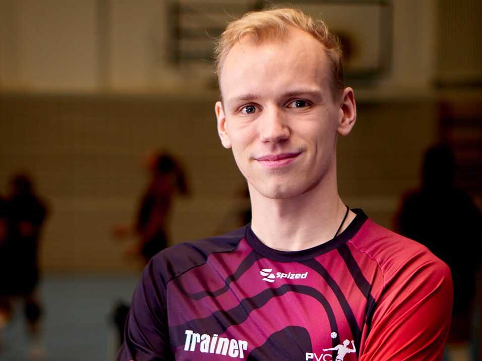 L&auml;chelnder Trainer in einem sportlichen Outfit mit &bdquo;PVC 91&ldquo;-Aufdruck, steht mit verschr&auml;nkten Armen auf einem Hallenboden.