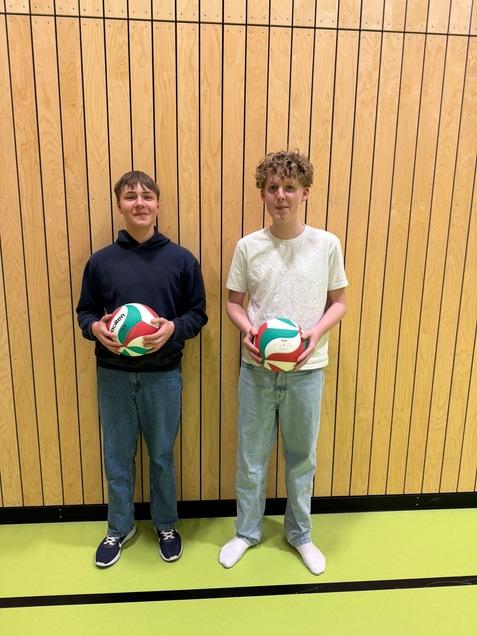 Zwei Jugendliche stehen mit Volleyballs vor einer Holzlattenwand in einem Sportraum. Einer trägt einen schwarzen Hoodie, der andere ein helles T-Shirt.
