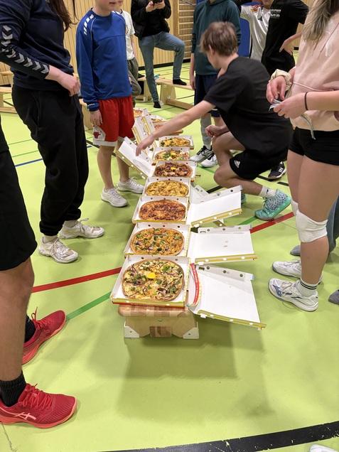 Gruppe von Jugendlichen um einen Tisch mit mehreren Pizzakartons in einer Sporthalle versammelt.