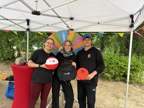 Drei Personen stehen unter einem Zelt, halten Frisbees und posieren vor einem Glücksrad. Hintergrund mit grünen Bäumen.
