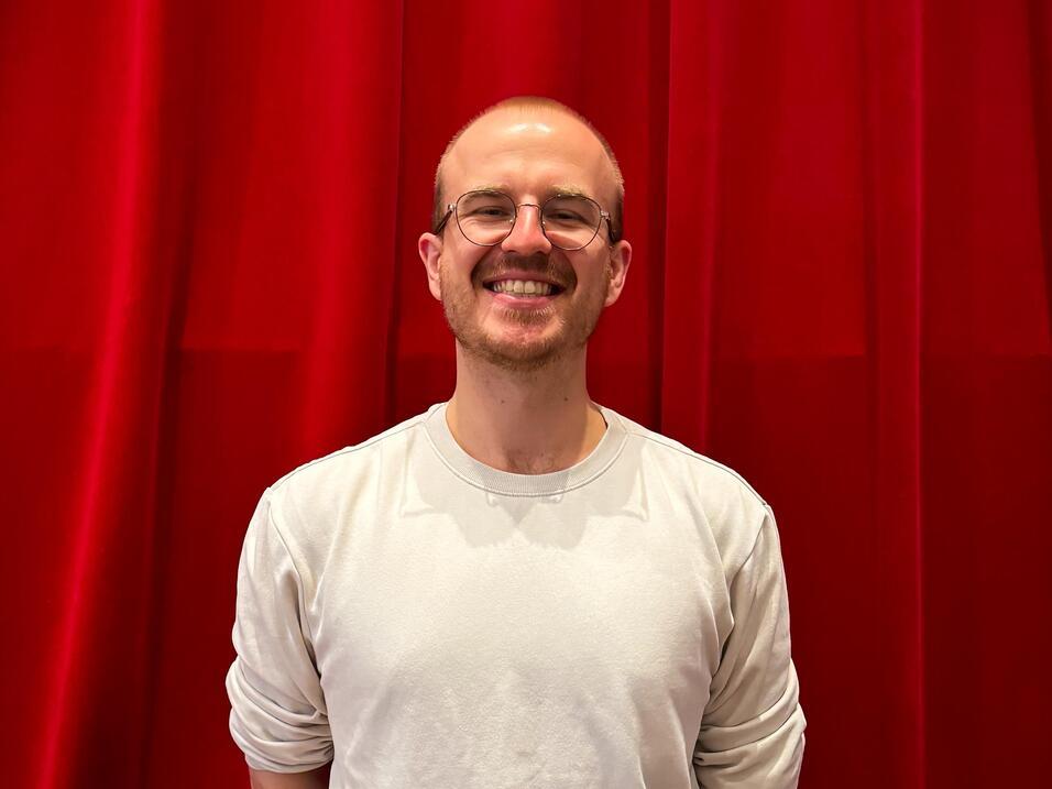 Mann mit Brille und kurzem Haar in einem weißen Shirt vor einem roten Vorhang.