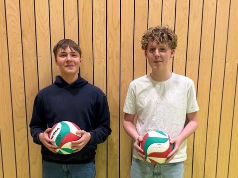Zwei Jugendliche stehen mit Volleyballs vor einer Holzlattenwand in einem Sportraum. Einer trägt einen schwarzen Hoodie, der andere ein helles T-Shirt.