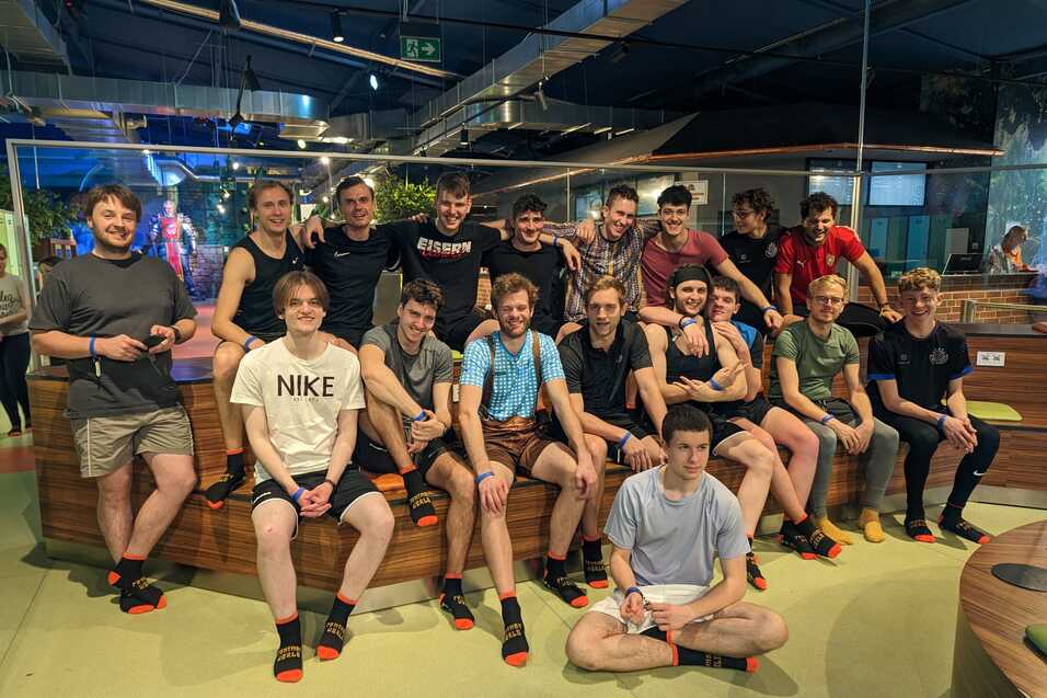 Gruppe von 16 jungen M&auml;nnern sitzt l&auml;chelnd auf einer Holzbank in einer Freizeitst&auml;tte mit bunten Socken.