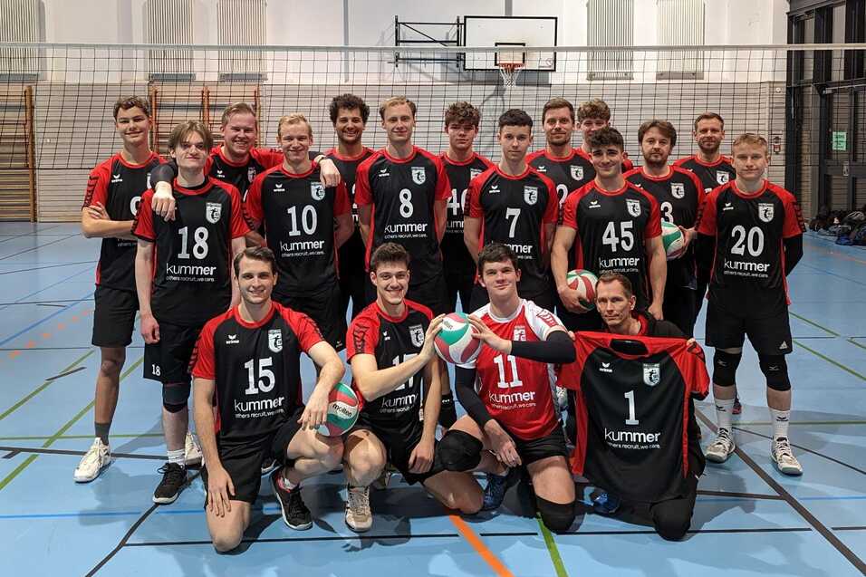Mannschaftsbild von 20 Volleyballspielern in roten und schwarzen Trikots, stehen in einer Sporthalle mit Netz und Bällen.