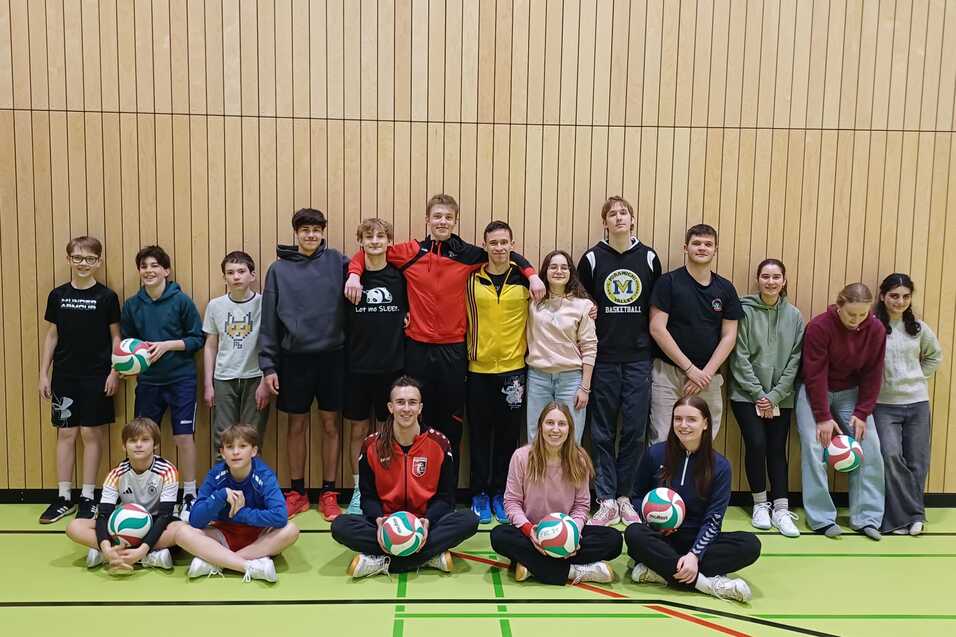Gruppe von Jugendlichen und jungen Erwachsenen mit Volleyballen in einer Sporthalle vor einer Holzpaneelwand.