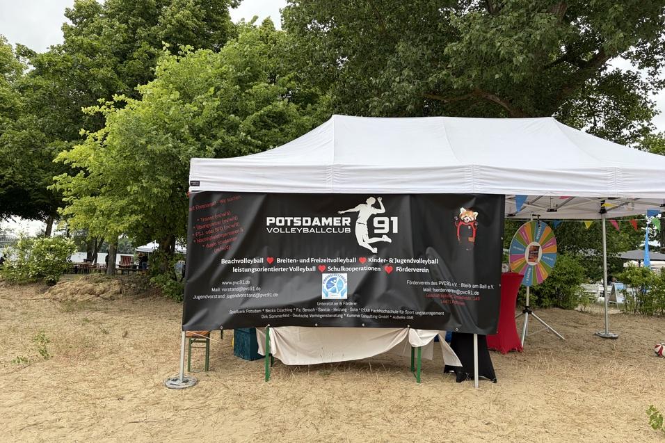 Zelt des Potsdamer Volleyballclubs mit Werbebanner und einem Glücksrad im Hintergrund. Sandboden und Bäume sichtbar.