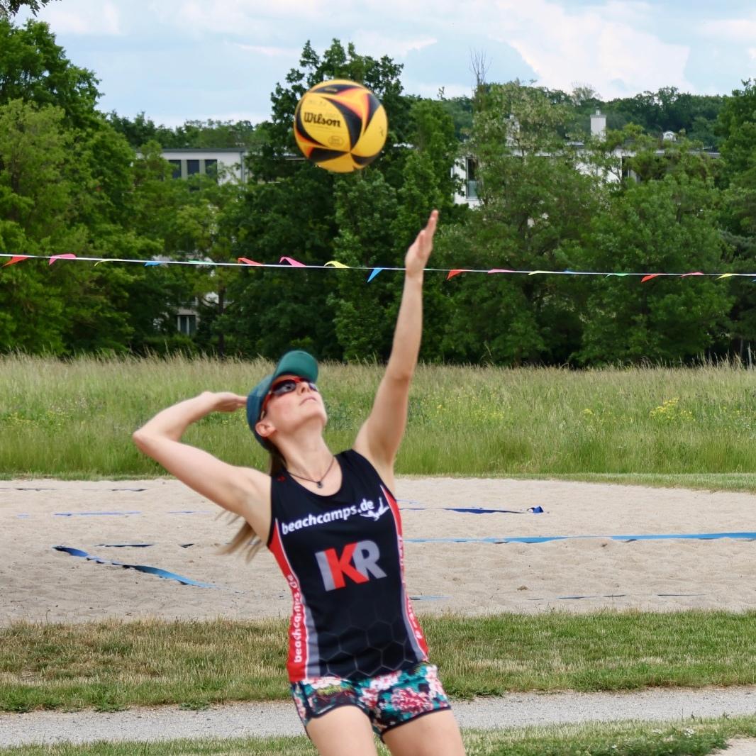 Sportlerin beim Volleyballspiel, hebt den Ball zum Aufschlag auf einem Sandplatz mit grüner Wiese im Hintergrund.