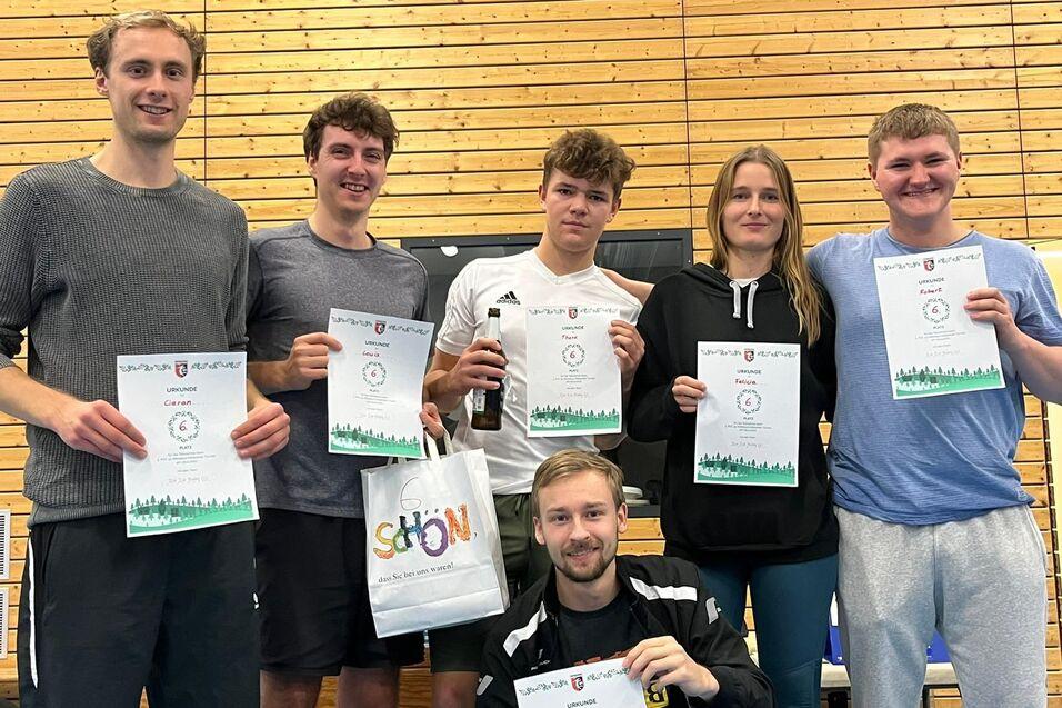 Gruppe von sechs Personen h&auml;lt Urkunden in die Kamera, umgeben von einer Sporthalle mit Holzpaneelen.