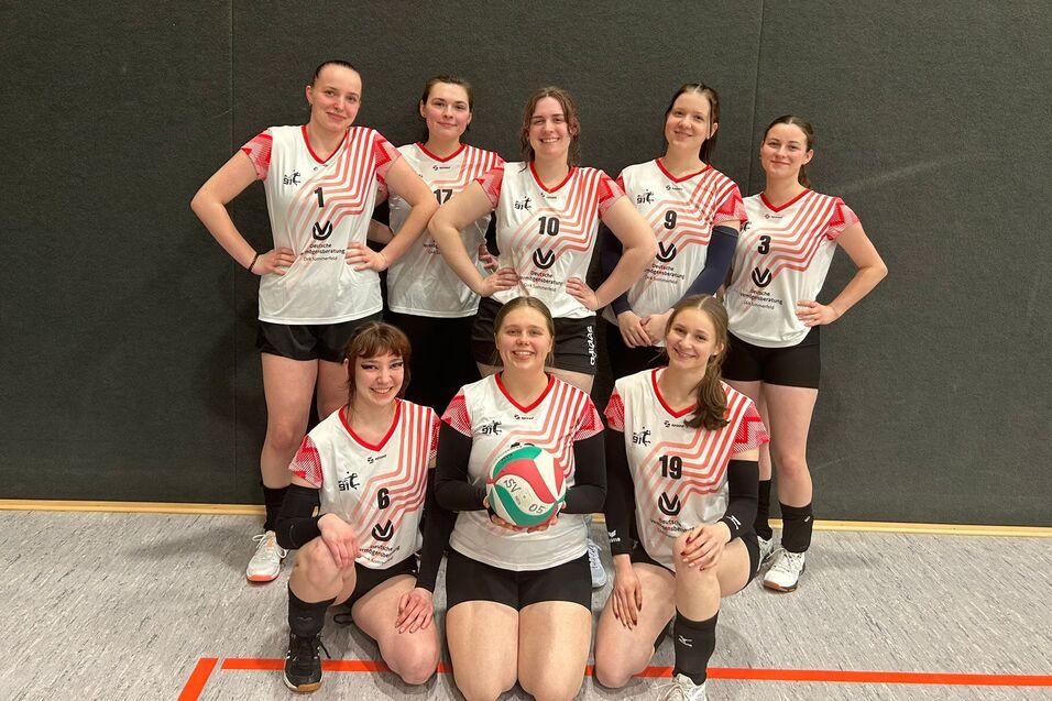 Acht weibliche Volleyballspielerinnen posieren in Sportkleidung mit einem Volleyball in der Mitte, vor einer grauen Wand.
