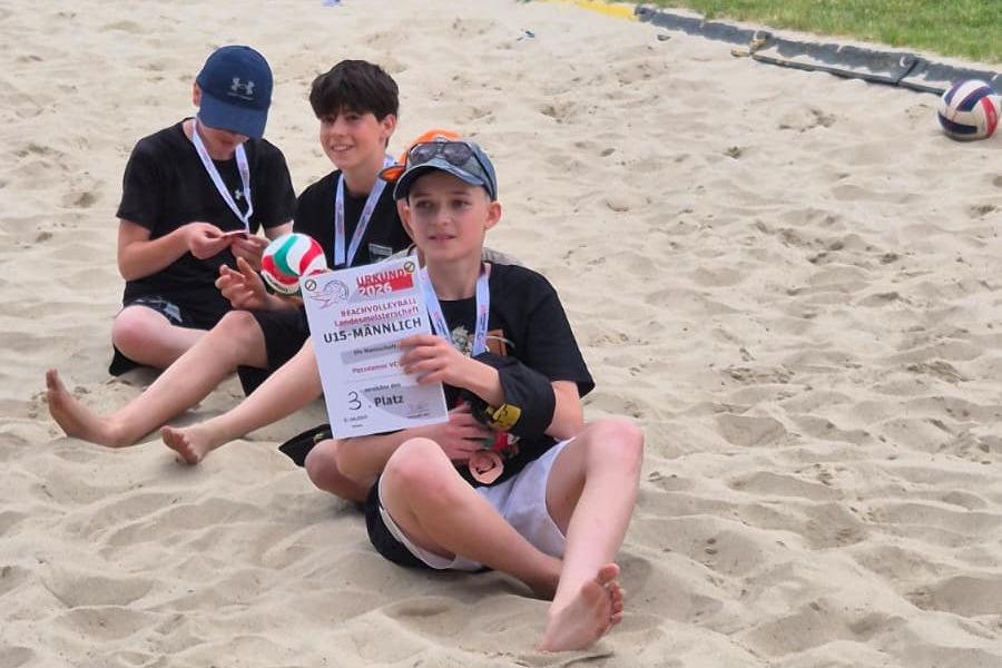 Drei Jugendliche sitzen im Sand, halten eine Medaille und eine Urkunde f&uuml;r den dritten Platz im Beachvolleyball.