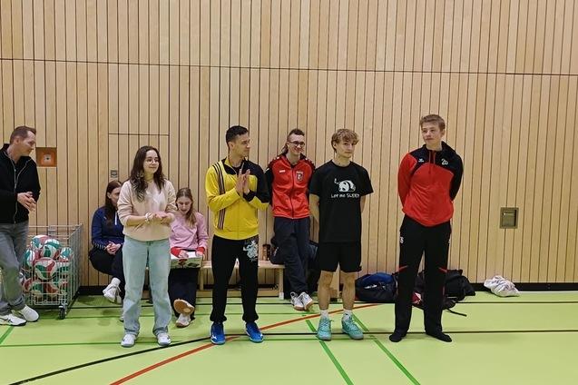 F&uuml;nf Personen stehen in Sportkleidung in einer Halle, w&auml;hrend im Hintergrund Zuschauer sitzen.