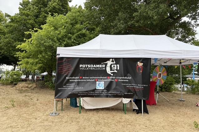 Zelt des Potsdamer Volleyballclubs mit Werbebanner und einem Gl&uuml;cksrad im Hintergrund. Sandboden und B&auml;ume sichtbar.