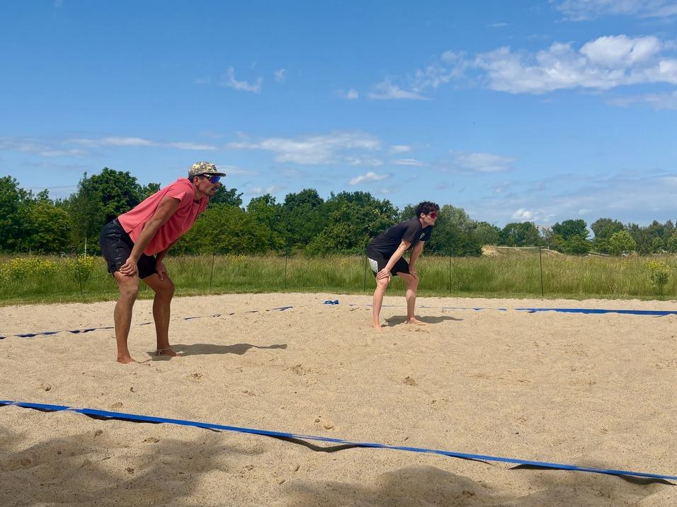 Zwei sportliche M&auml;nner in Umkleidung stehen auf einem Sandplatz, bereit f&uuml;r eine Sportaktivit&auml;t unter blauem Himmel.