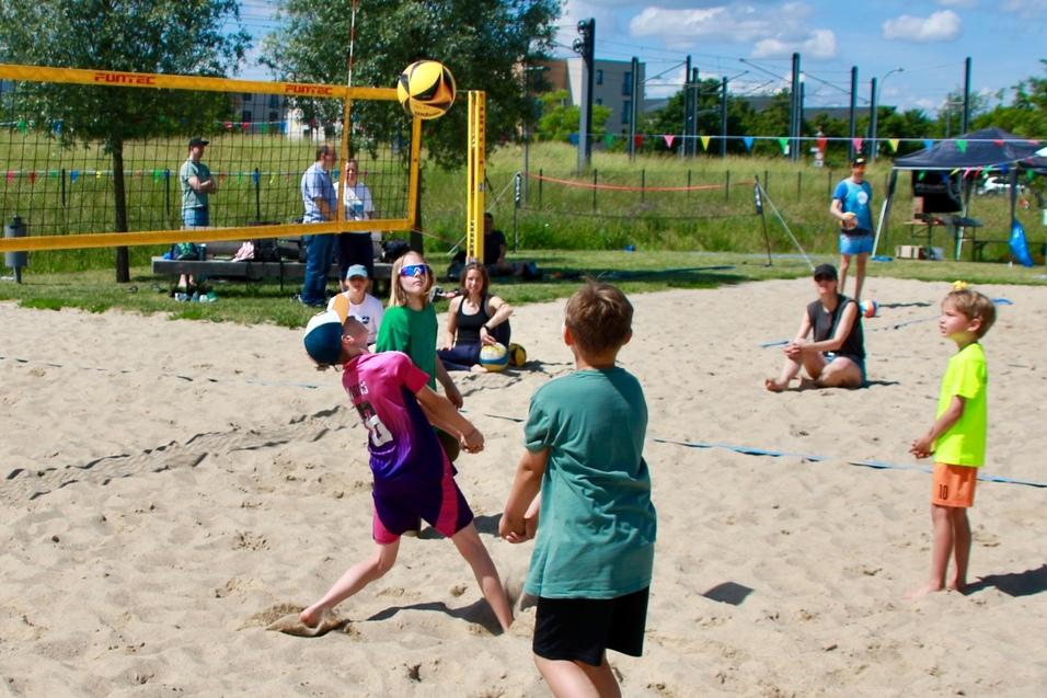 Vier Kinder spielen Beachvolleyball im Sand, w&auml;hrend Zuschauer im Hintergrund stehen und zuschauen.