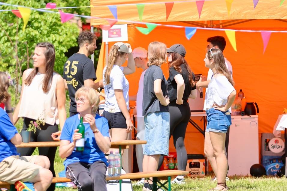 Gruppen von Menschen stehen an einem orangefarbenen Zelt, darunter Tischtennisutensilien und Getränke auf einem Tisch.