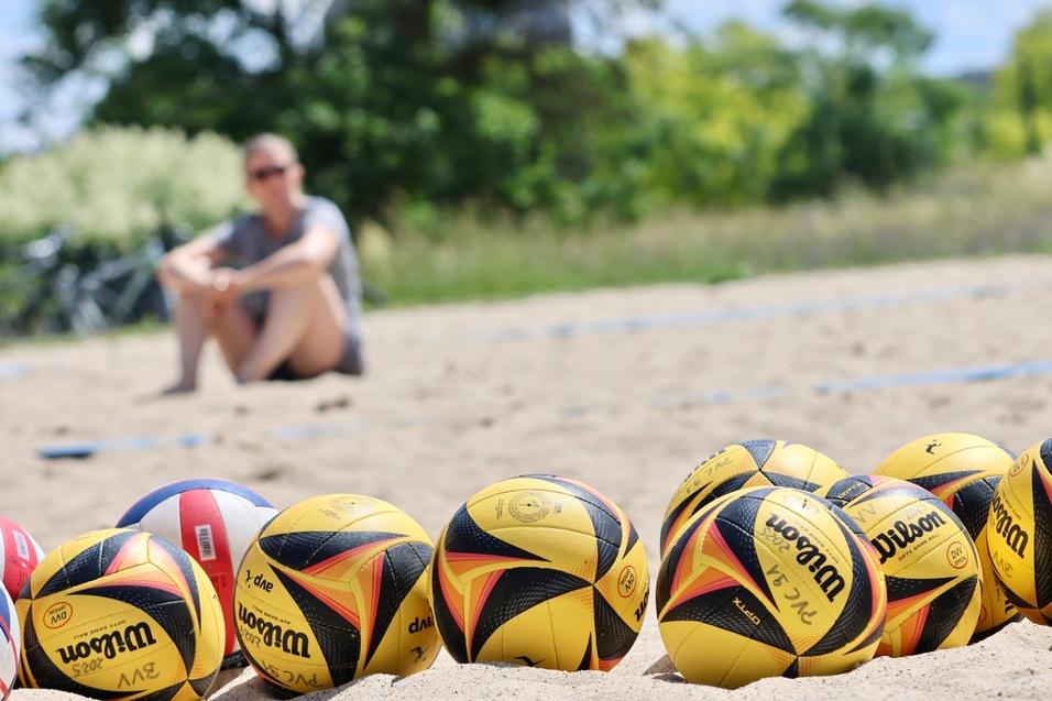 Reihe von gelben Beachvolleyballen mit schwarzem Logo auf sandigem Untergrund, im Hintergrund eine Person sitzend.