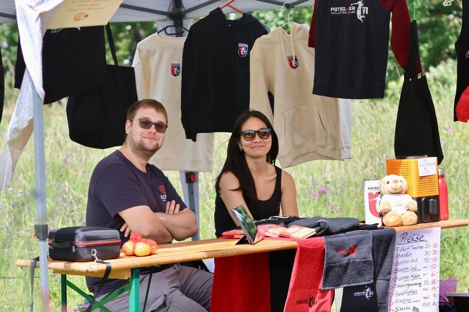 Zwei Personen sitzen an einem Marktstand mit Bekleidung und einem Schild mit Preisen, umgeben von gr&uuml;ner Wiese.