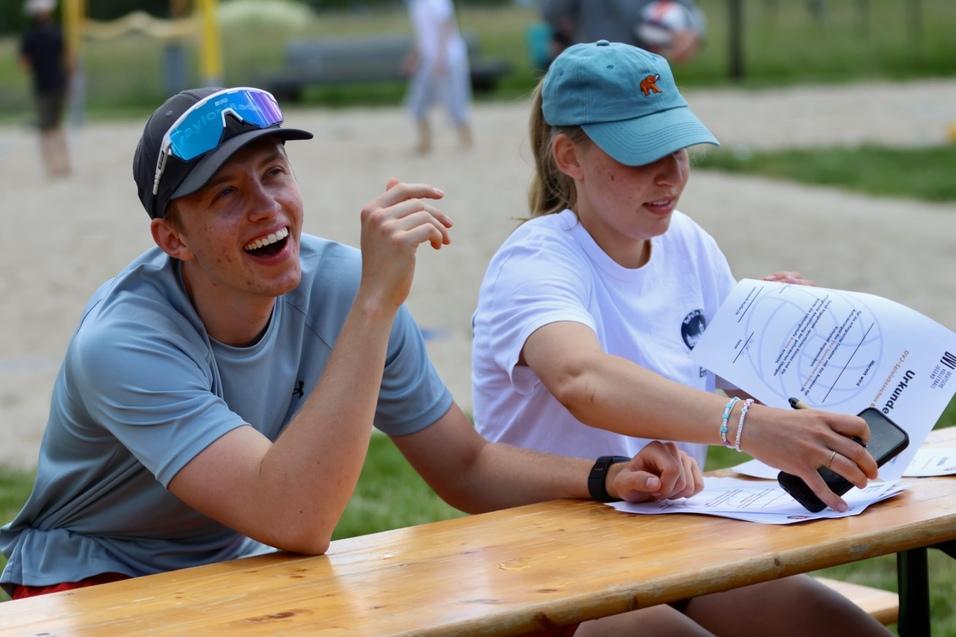 Zwei Personen sitzen an einem Tisch, lachen und halten Papiere, im Hintergrund ein Sandplatz und eine Spielger&auml;teecke.