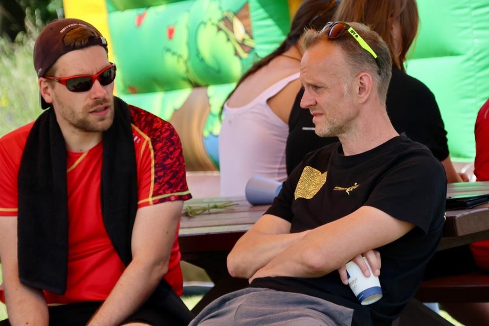 Zwei M&auml;nner sitzen an einem Tisch, einer tr&auml;gt ein rotes Shirt, der andere ein schwarzes, im Hintergrund ist ein H&uuml;pfburg sichtbar.