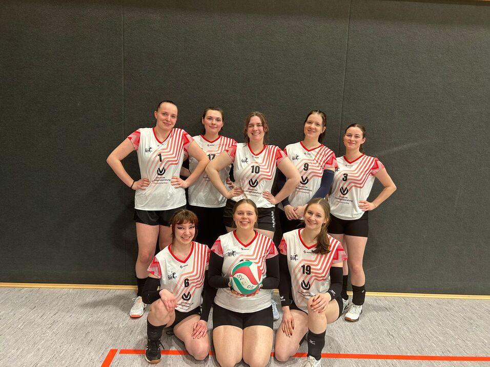 Acht weibliche Volleyballspielerinnen posieren in Sportkleidung mit einem Volleyball in der Mitte, vor einer grauen Wand.