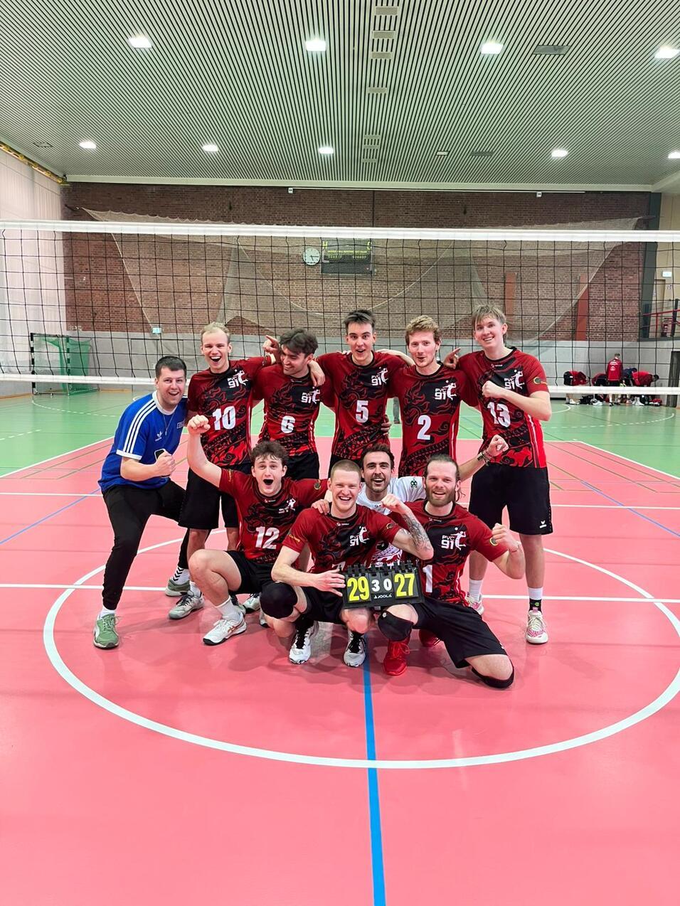 Gruppe von Volleyballspielern in rot-schwarzen Trikots posiert freudig auf dem Spielfeld nach einem Sieg.