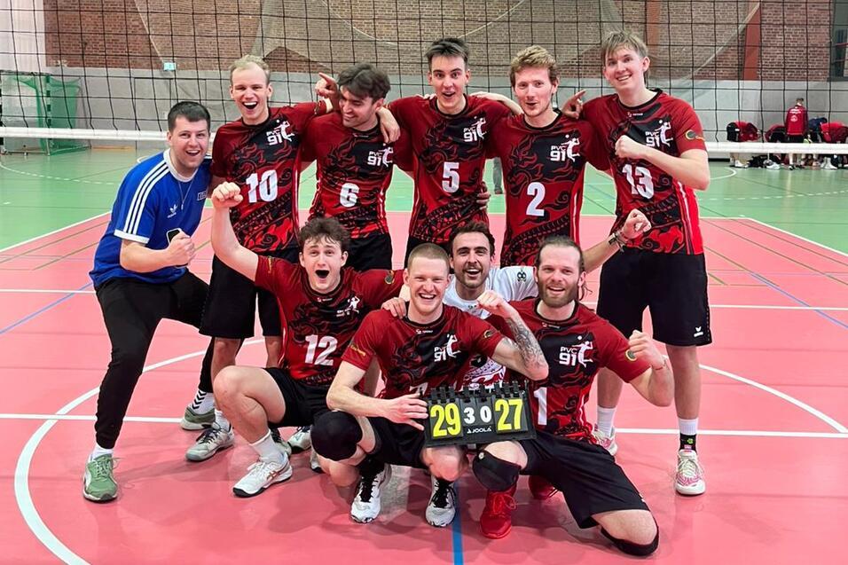 Gruppe von Volleyballspielern in rot-schwarzen Trikots posiert freudig auf dem Spielfeld nach einem Sieg.