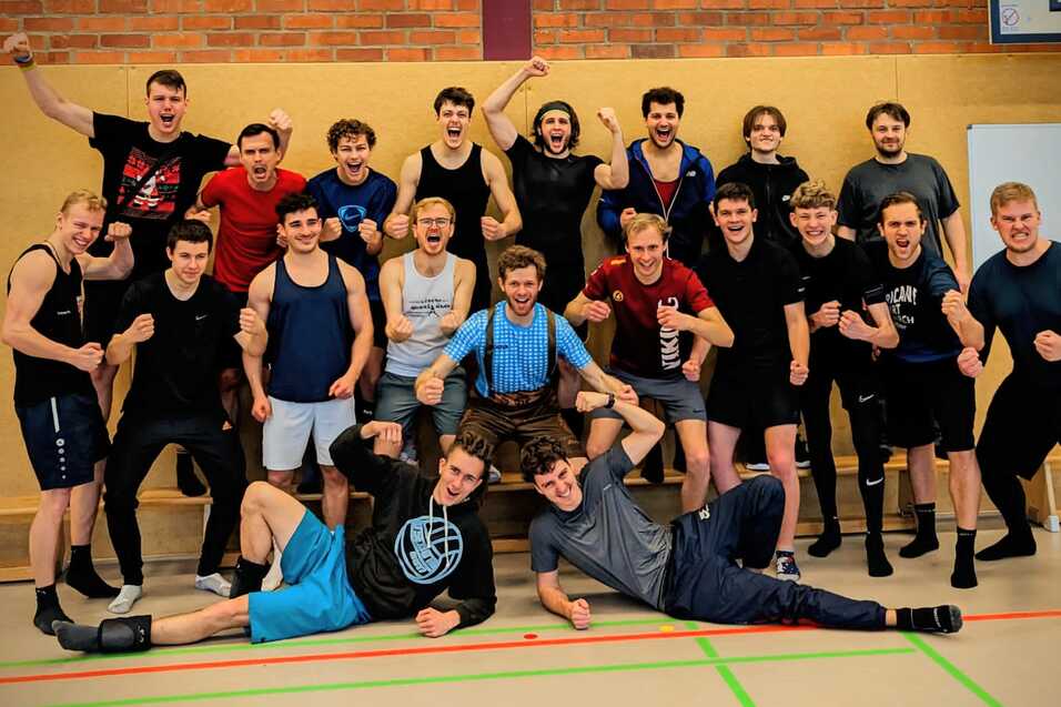 Gruppenfoto von 20 m&auml;nnlichen Ringertrainierenden in Sportkleidung, die in einer Sporthalle posieren und jubeln.