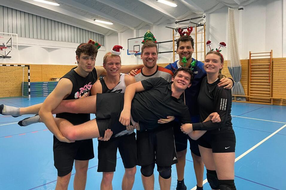 Sechs sportlich gekleidete Personen posieren in einer Sporthalle, zwei tragen lustige Weihnachtsaccessoires.